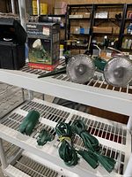 Photo showing two round ground landscape floodlights and the boxed Woods Floodlight Kit transformer on a white wire shelf.