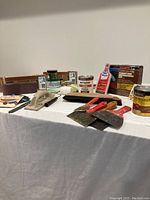 Photo of painting and refinishing supplies including sanding block, putty knives, Rust-Oleum Metal Primer, Minwax Wood Finish, furniture refinishing kit, brushes, and paint brush cover arranged on a white tablecloth.