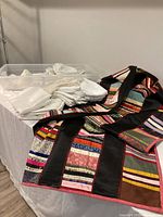 Quilted patchwork spread with colorful fabric strips separated by black sashing, alongside stacks of white half-window curtains.