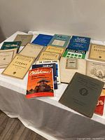 Overview of 18 sheet music pieces laid out on a white table showing a variety of classical and popular music titles.