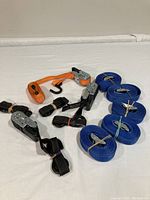 Assortment of seven ratcheting tie down straps on white cloth background, including four blue, two black, and one orange strap, all rolled and bundled with rubber bands.