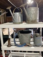 Photo of two watering cans on top shelf, and a metal pail, gas can, and oil can on a lower shelf in a storage setting.