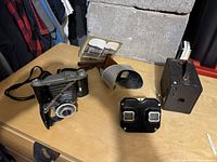 Four antique photography items displayed on a wooden table including three vintage cameras and a vintage stereo viewer with stereograph cards.