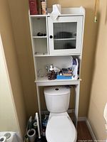 White over-the-toilet bathroom shelf cabinet with compartments and glass door, shown in a bathroom space with a toilet underneath and some items stored on shelves.