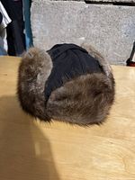View of the black wool winter dress hat with natural brown muskrat fur trim from the side angle on a wooden surface.
