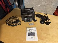 Dash camera with lens, backup camera, cables and mounts, user manual, and hardware on wooden table with product box in background.