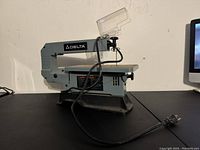 Front angle view of the Delta scroll saw on a black surface showing the work table, arm, and power cord.