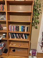 Front view of a tall light brown wooden bookshelf with four shelves containing various books and small miscellaneous items.