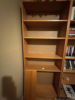 Photo showing the front view of the tall light brown bookshelf with multiple empty shelves and a wider lower compartment.