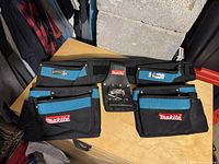 Front view of Makita tool belt showing black and blue color scheme, multiple pockets including two large front pouches, and the Makita logo clearly visible. Velcro waist strap is seen attached with additional belt loops.