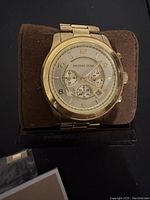 Front view of gold tone Michael Kors chronograph wristwatch with subdials on a brown cushion and holder.