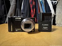Front-facing Panasonic LUMIX digital camera with lens retracted, showing Leica lens branding and 16x optical zoom text, alongside the black Panasonic battery charger.