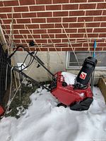 Side view of red Toro snow thrower with black chute and handle in snow next to brick wall.