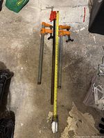 Three bar clamps arranged on the floor, two 24-inch clamps with orange handles and one 36-inch clamp with a red handle and measuring tape alongside for scale.