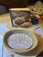 Photo showing artisan bread cookbook standing upright behind a ceramic bread warmer with tree embossing inside a woven basket on a wooden table with a coffee mug nearby.