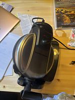 Hamilton Beach waffle maker atop wooden table with surrounding papers and a book. Stainless steel body with black handle and control dial visible.