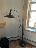 Full view of industrial style floor lamp with adjustable arm and wide conical metal shade near window.