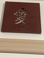 Wooden square plaque with metal cutout character for 'love' and English text 'love' below, reddish-brown color.