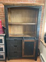 Front view of black wooden kitchen hutch showing two open shelves at top, three drawers in middle, and sliding door shelf on right