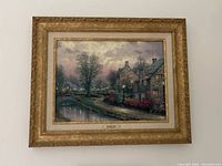 Photo of framed Lamplight Lane by Thomas Kincade hanging on wall, showing scene with houses, trees, bridge and waterway at dusk in ornate gold frame.