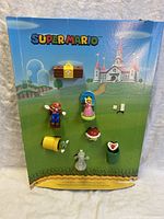 Front view of Super Mario-themed McDonalds Happy Meal toy display board with seven toys mounted on a colorful background showing Princess Peach, Mario, pipes, and castle.