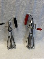 Two vintage handheld manual kitchen beaters side by side showing metal frame, dual rotating beaters, and crank handles with red and black molded plastic handles.