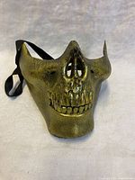Front view of gold skull masquerade mask on white background, showing detailed teeth, nose, and eye cut-outs.