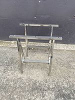 Pair of chrome A frame saw horses standing on pavement against a black wall, showing full front and side profile.