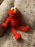 Full view of the red 24-inch Sesame Street Tickle-me Elmo plush toy sitting on a floral patterned couch.