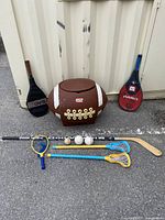 Photo showing large football-shaped toy box flanked by two racquets (Head and Wilson) and arranged items including lacrosse sticks, balls, and small baseball bat