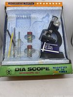 Top-down view of the full microscope set showing all included items in the box including microscope, slides, dropper, containers, tweezers, and brush