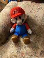 Mario plush sitting on a patterned beige armchair, visible from front with clear detail of the hat emblem, eyes, and overalls buttons