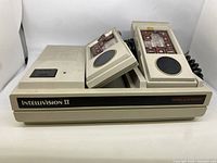 Front side view of beige Mattel Intellivision II console showing integrated slot and two connected controllers with round directional pads and button overlays.
