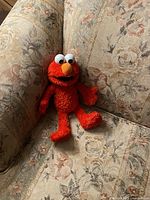 Full-body view of the red Elmo plush sitting on a floral patterned couch, showing its fur texture and sitting pose.