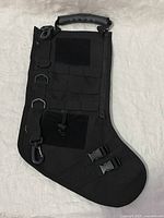 Front view of the black tactical Christmas stocking showing MOLLE straps with carabiners, quick-release clips on the toe, hook-and-loop panel, zipper, and handle.