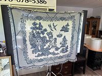 Afghan throw blanket hanging displaying entire patterned surface with blue and white scenic design including birds, trees and pagodas.