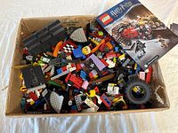 Photo showing box with various loose LEGO pieces including bricks, wheels, and minifigure parts alongside LEGO Harry Potter instruction manual.