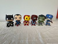 Front view of six Marvel/DC Funko Pop figures on white fabric, showing Batman, Superman, Iron Man, Spider-Man, Hulk, and Captain America.