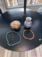 Top-down image of the round black table displaying the two cloisonné necklaces, decorative round box, and blue and white porcelain vase.