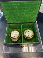 Pair of two vintage brass automotive dash clocks in green velvet lined wooden box; left is diamond-shaped Dieppe clock, right is round Ormond clock with cracked glass.