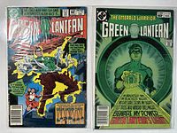 Two Green Lantern comic books in protective plastic sleeves, front covers visible. One cover features a battle scene with Green Lantern, the other features Green Lantern holding a glowing lantern symbol.