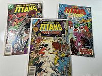 Three vintage Teen Titans comic books shown in plastic sleeves on white surface, visible titles include Tales of the Teen Titans and The New Teen Titans.