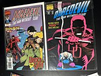 Two Daredevil comic books showing distinct covers: one with Daredevil protecting a woman and another with large pink/black figure behind kneeling Daredevil