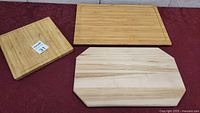 Three wooden cutting boards including a large rectangle with juice groove, square board, and unique angled corner board on red surface.