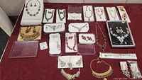 Overview of assortment of costume jewelry sets on a red draped surface. Includes multiple necklaces and earrings in varying designs and colors such as gold-tone red rhinestones, black and gold clusters, and blue rhinestones.