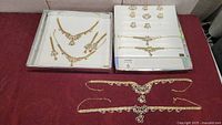 Three sets of costume jewelry including necklaces, bracelets, and earrings displayed in trays and on table.