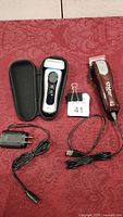Set of a Braun electric shaver with its protective case and charger plus a brown Wahl hair clipper with charging cable and charger on red patterned fabric.