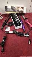 Overview of four hair styling tools on red cloth: boxed professional hair straightening brush, two hair straighteners, two curling irons including purple and pink spiral style.