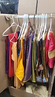 Photo showing nine garments including sarees and dupattas hung on plastic and wooden hangers in a cardboard box. Colors include red, peach, yellow, olive green, purple, pink. Variety of fabrics and textures visible.