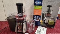 Oster Jus Simple Juicer with original box, pulp container, juice cup, and instruction manual on a red cloth surface.